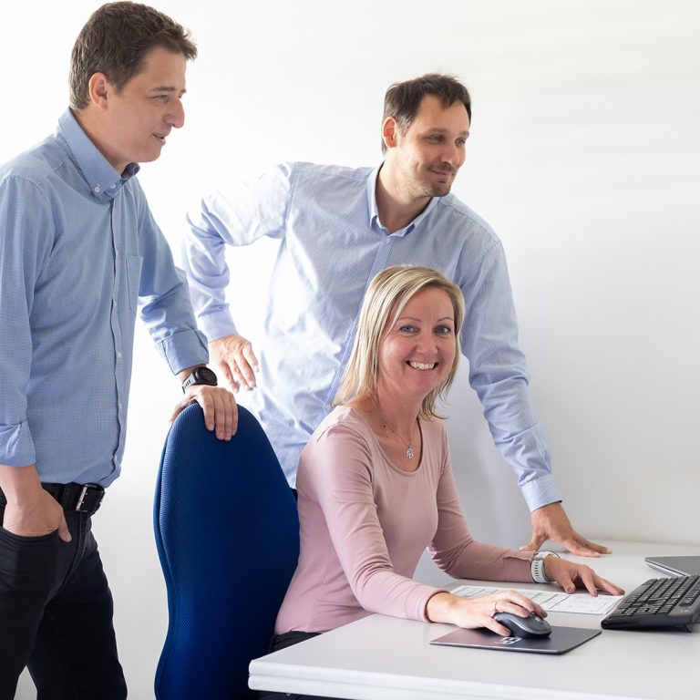 The picture shows three employees sitting in front of a computer in the project management office.